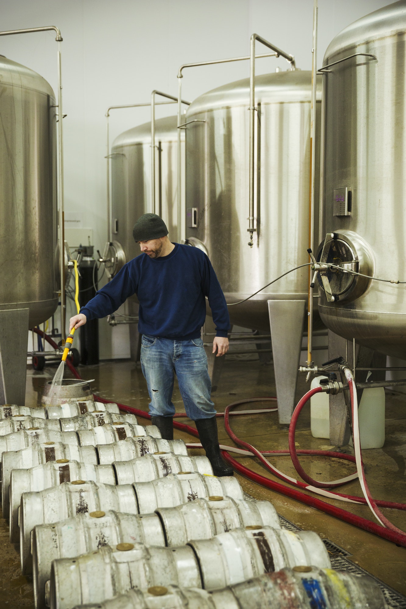 Man standing beside the fermentation tanks, cleaning metal beer kegs with water from a hosepipe.